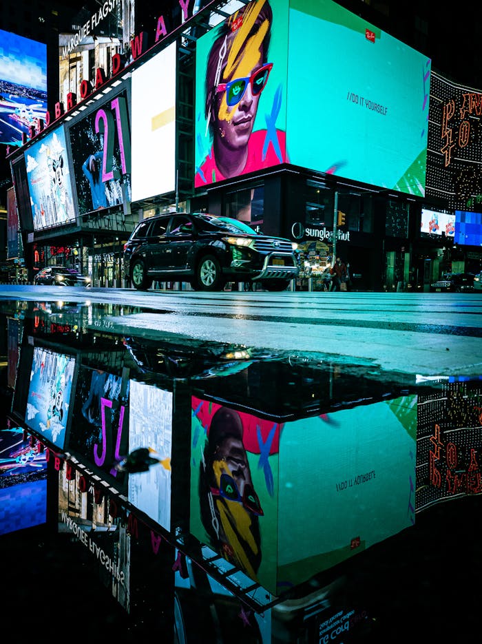 A dynamic scene of illuminated billboards reflected in a puddle on a bustling city street at night.